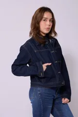 Campera de jean de color azul oscuro, con corte cropped y costuras en contraste color caramelo. Presenta cuello clásico, cierre frontal con botones metálicos y bolsillos a la altura del pecho y laterales.