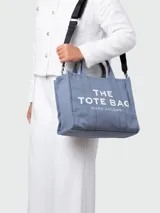 Bolso tote mediano de lona color celeste con estampado del logo "The Tote Bag" en blanco y nombre de la marca "Marc Jacobs" en blanco. Incluye doble asa corta y correa larga extraíble y ajustable en color negro.