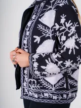 Chaqueta corta negra con bordado floral blanco, cuello camisero y mangas largas.