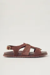 Sandalia plana de gamuza color marrón oscuro, con tiras cruzadas en la parte delantera y una tira fina ajustable con hebilla metálica en el tobillo. Posee plantilla de cuero y suela baja.