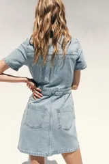 Vestido corto de denim azul, con cuello solapa, escote en V, manga corta, bolsillos delanteros y cierre frontal con botones metálicos.