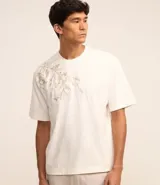 Remera de algodón color crema, corte boxy, cuello redondo y manga corta. Presenta un bordado floral en el lateral superior izquierdo.