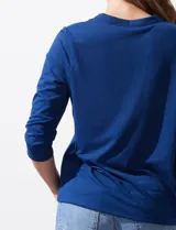 Blusa blanca de poliéster y elastano, con cuello a la base, escote en V y mangas tres cuartos. Presenta dos bolsillos frontales con cierre metálico dorado.