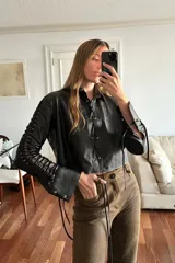 Camisa corta de cuero ovino negro con cuello clásico, cierre frontal con cordones y mangas largas con aberturas geométricas.