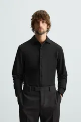 Camisa color beige de corte slim fit, confeccionada en tejido elástico que minimiza las arrugas. Tiene cuello italiano abierto, manga larga con puños abotonados y cierre frontal con botones.