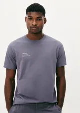 Camiseta blanca de algodón con cuello redondo y mangas cortas. Estampa rectangular con borde negro que contiene la frase "NATURAL ATMOSPHERE" en letras mayúsculas y una imagen de montañas y mar.
