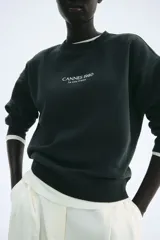 Buzo de tejido de punto color verde oscuro, con cuello redondo, hombros caídos y puños y bajo acanalados. Presenta un pequeño estampado centrado en el pecho con el texto "CANNES 1980" y debajo "La Côte D'Azur".