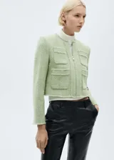 Chaqueta corta color verde claro de tweed con cuello redondo y cierre frontal con cremallera. Tiene cuatro bolsillos de parche en la parte delantera y mangas largas.
