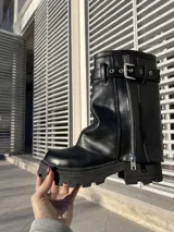 Botas negras de ecocuero con caña media, cartera con cierre y cinto decorativo.