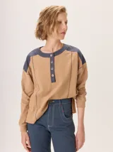 Buzo de manga larga con diseño de bloques de color y textura. El cuerpo principal es marrón, la espalda superior y hombros tienen un panel de tejido a rayas grises y azules, y los hombros superiores son de color beige rosado. Presenta costuras decorativas en rojo que separan los bloques de color.