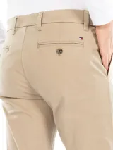 Pantalón chino color beige de corte slim, con cierre de botón y cremallera, cuatro bolsillos y tejido elástico. Cuenta con detalles distintivos de Tommy Hilfiger y bandera de la marca en la parte posterior.