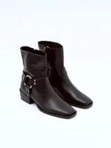 Bota de cuero negra con caña media, puntera ligeramente afilada y detalle de correa con herraje metálico circular y tachas. Taco bajo y ancho.