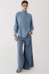 Camisa celeste de jean con cuello alto con volados y mangas largas abullonadas con puños con volados. Presenta botones de madera y costuras rojas a contraste.