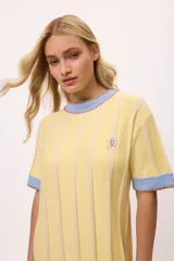 Vestido amarillo de algodón con rayas verticales en tono crema, cuello redondo y mangas cortas con ribete celeste y bordes en marrón. Presenta un logo bordado en el pecho.