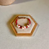 Pulsera elástica con cuentas de cerámica blanca decoradas con motivos florales rojos y marrones, separadas por pequeñas cuentas rojas. Cierre de mosquetón dorado y cadena de eslabones dorada.