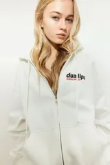 Campera blanca con capucha y cierre frontal, con bolsillos tipo canguro y puños acanalados. Estampado frontal con el logo de Dua Lipa y la frase "Training Season".