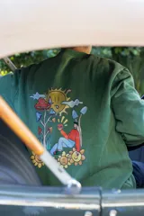 Buzo verde con estampa en la espalda de un sol, nubes, hongos y un personaje sentado sobre flores con una hamburguesa en la mano.