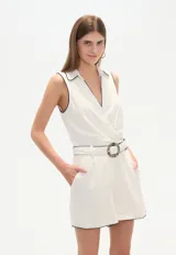 Mono corto color blanco, sin mangas, con cuello camisero, corte cruzado y cinturón con hebilla circular.