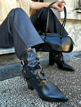 Botas de cuero sintético negro con punta fina, caña media con correas y hebillas plateadas, y taco cuadrado.