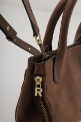 Cartera pequeña de cuero marrón oscuro, con asas superiores y cierre de cremallera lateral visible. Presenta el logo de la marca grabado en el frente.