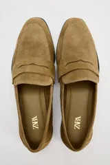 Zapato plano tipo mocasín color marrón claro, confeccionado en piel de serraje. Tiene copete subido, antifaz y punta redonda.