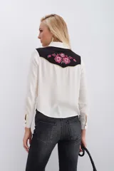 Camisa blanca de manga larga con canesú delantero y trasero en tela negra con bordados florales en tonos rosados y detalles en hilo dorado. Cierre frontal con botones metálicos tipo snap.