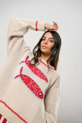 Sweater tejido color crudo con cuello redondo y mangas largas. Presenta dos peces rojos con detalles en azul bordados en el frente. Tiene ribete ondulado rojo en cuello, puños y ruedo.