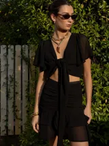 Conjunto de top y falda negros. El top es corto, con escote en V, mangas cortas amplias y lazo en el frente. La falda es corta, con frunces y volados.