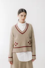 Sweater beige con cuello en V y mangas largas con diseño trenzado. Tiene dos bolsillos frontales con bordados de tréboles rojos y ribetes rojos en cuello, puños y ruedo.