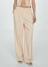 Pantalón de vestir color beige, de tiro alto, corte wide leg, con cintura elástica, pliegues frontales, cierre de cremallera y botón, y bolsillos laterales.