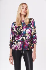 Camisa estampada con flores en tonos morados, azules, amarillos y verdes sobre fondo negro. Tiene cuello redondo, mangas abullonadas 3/4 y lazo en la cintura.