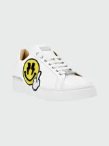 Zapatillas bajas de cuero blanco con aplique de parche bordado de cara sonriente amarilla con relámpagos en los ojos y dedo medio levantado.