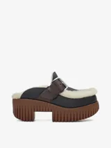 Zapato tipo mule con plataforma track de color marrón y capellada de cuero negro. Cuenta con forro de lana de oveja (borreguito) visible en los bordes e interior, y una correa de cuero marrón con hebilla metálica sobre el empeine.