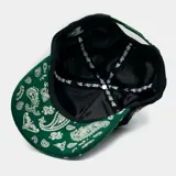 Gorra de béisbol negra con visera estampada con diseño paisley verde y blanco. Presenta un bordado de la Virgen de Guadalupe en el panel lateral y una letra 'M' estilizada en el frente con franjas blancas, verdes y doradas.