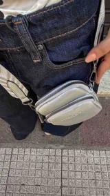 Bandolera pequeña tipo 'phone bag' de símil cuero color rosado pálido, con cierre de cremallera y correa ancha de tela con diseño de chevron en tonos claros.