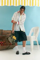 Camisa blanca oversize de algodón con bordado calado tipo broderie, cuello camisero con ribete rojo, cierre frontal con botones y mangas largas con puños con ribete rojo. Presenta una franja vertical con ribete celeste y otra con ribete amarillo.