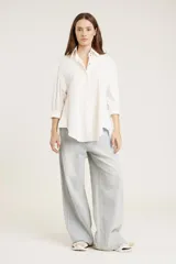 Camisa blanca de corte clásico con cuello abotonado, cierre frontal con botones y mangas largas con puños abotonados. Presenta pequeños apliques de strass plateados dispersos por toda la prenda.