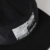 Gorro tipo trucker negro con paneles frontales de tela y paneles traseros de malla. Presenta un parche frontal blanco con un diseño gráfico abstracto dividido en cuatro secciones y el texto "FORMER MERCH" debajo.