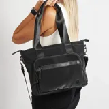Bolso tote negro con doble asa de hombro y correa bandolera extraíble. Tiene un bolsillo frontal con cierre y un logo circular en la esquina inferior.