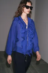 Camisa azul con escote en pico, cierre frontal con botones, mangas largas abullonadas con elástico en los puños y detalle de volantes en el frente.