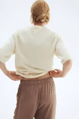 Remera de punto color crema, de corte holgado con hombros caídos y mangas cortas al codo. Presenta cuello redondo y remates acanalados en cuello, puños y bajo.