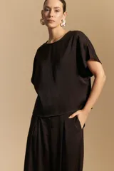 Top negro de manga corta con silueta amplia y caída fluida, confeccionado en un tejido liviano con leve brillo. Presenta un botón de nácar en la espalda con una pequeña abertura y mangas amplias tipo kimono.