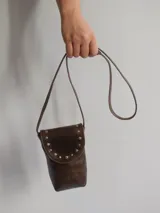 Bandolera porta celular marrón de cuero con solapa y cierre con botón a presión. Presenta tachas metálicas plateadas en el borde de la solapa y correa fina para colgar al hombro.