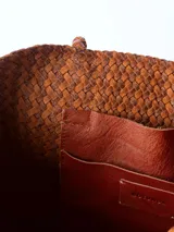 Bolso tipo tote bag color marrón, confeccionado en cuero trenzado. Tiene doble asa de hombro y base rectangular.