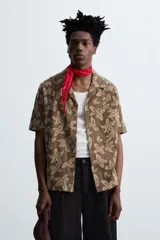 Camisa marrón con estampado floral, corte relaxed fit, cuello bowling, manga corta, bolsillo en el pecho, aberturas laterales y cierre con botones.