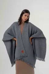 Ruana de punto color gris con diseño de trama diagonal y terminación de flecos en el ruedo. Presenta un pequeño parche decorativo en la parte frontal inferior.