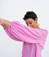 Blusa tipo blusón color rosado, de viscosa, con cuello redondo, manga larga acampanada, pliegues en los hombros, detalle de entredós en el escote y tiras de ajuste con borla.