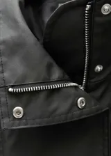 Anorak negro oversize con cuello alto, cierre frontal con botones y cremallera oculta, dos bolsillos laterales con cremallera, mangas largas con puños abotonados y charreteras en los hombros.