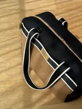 Bolso tipo tote de neopreno color negro y marrón, con cierre superior, bolsillo frontal marrón con logo rojo, bolsillo interno y externo con cierre, doble asa de mano marrón con detalles blancos y opción de correa larga.