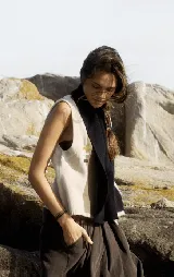 Chaleco de tejido de punto sin mangas, con diseño de bloques de color verticales en tonos beige claro, marrón topo y negro. Presenta un cuello chal negro y tres pequeños bordados horizontales en amarillo, lila y marrón en el frente.